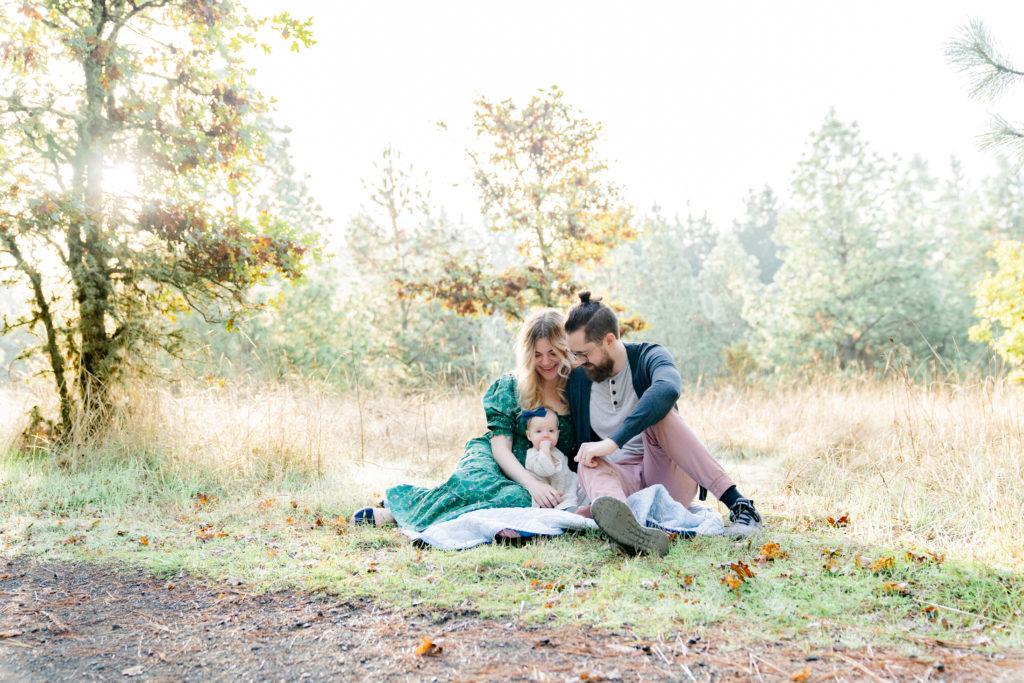 photo of portland oregon family of three sitting in a field surrounded by trees with the sun pouring around them